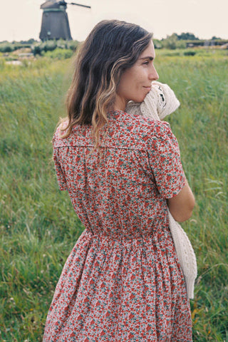 From behind, a woman wearing a red and green apple print short-sleeve dress stands in a grassy field with a windmill and cloudy sky in the background, holding a cream knit sweater.