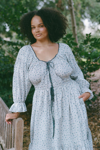 Woman wearing a light blue floral dress standing outdoors with greenery in the background