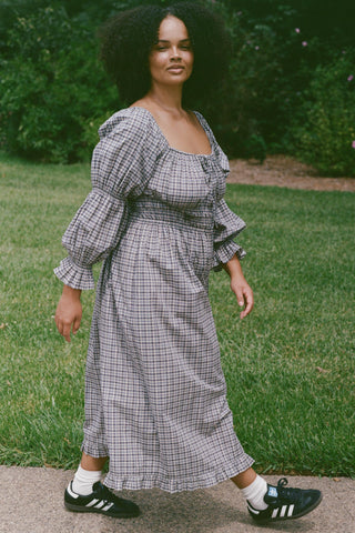 Woman wearing a plaid dress with Adidas shoes walking on a stone path with grass and bushes behind her