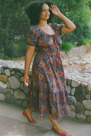 Woman wearing a patterned dress walking outdoors near a stone wall.
