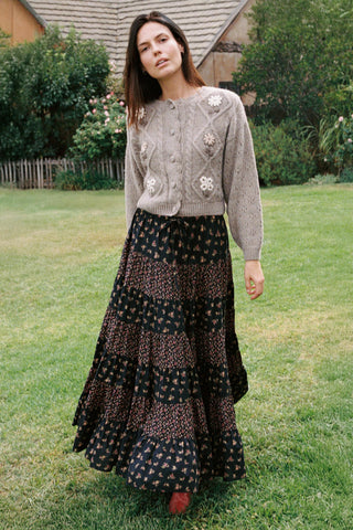A woman stands in grass, wearing a patterned cardigan, a black flowing skirt, and red socks.