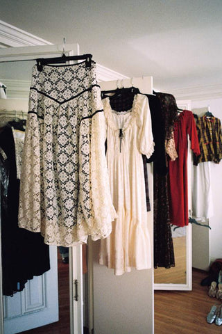 A black and white patterned lace dress hanging on a clothes rack in a room, with other dresses in cream, black, and red hanging behind it.