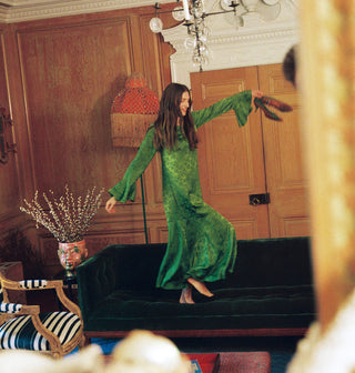 A woman in a long green dress dances on a green velvet couch while holding her shoes