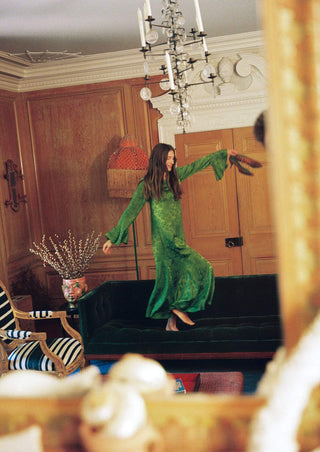 A woman in a flowing green patterned dress stands barefoot on a dark green velvet sofa in an ornate room with wood-paneled walls and a chandelier.