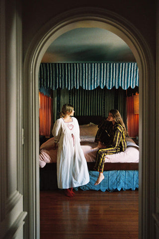 A woman in a white nightgown stands beside another woman in a green and gold patterned pajama set on a bed in a dimly lit bedroom, viewed through an arched doorway.