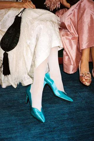 A close-up lifestyle shot of a woman wearing shiny turquoise pointed-toe high heels with white tights, seated next to others in formal attire on a dark blue carpet.