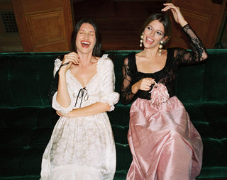 Two women laughing and posing together on a dark green velvet sofa. The woman on the left wears a white lace dress with black trim, and the woman on the right wears a black lace top with a pink satin skirt.