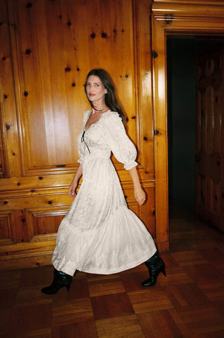 A woman wearing a long white puff-sleeve midi dress with black ankle boots walks through a room with warm wood-paneled walls and parquet flooring.