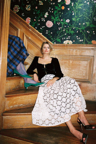 A woman sits on wooden stairs wearing a black velvet long-sleeve top and a white lace midi skirt with a black geometric pattern. She is posed against a background of dark floral wallpaper and wood paneling.