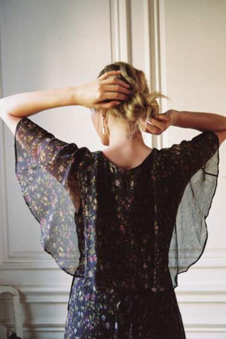 A woman viewed from behind wearing a sheer black floral blouse with wide flutter sleeves, standing indoors against a light paneled wall.