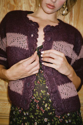 A close-up of a woman wearing a dark plum purple and dusty pink striped knit cardigan over a black floral dress, standing against a light wood-paneled wall.