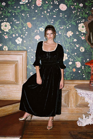 A woman models a black velvet midi dress with a square neckline, ruffled sleeves, and a gathered waist, standing in a room with a dark floral wallpaper background.