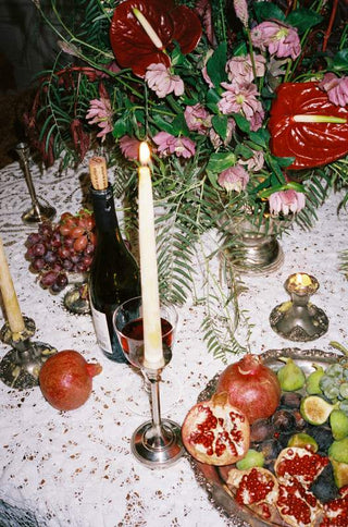 A styled tabletop scene with a floral arrangement, lit taper candles, a dark wine bottle, grapes, pomegranates, figs, and a platter of fruit on a white patterned tablecloth.