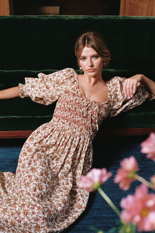A woman wearing a knee-length floral dress with a tie neckline, posed on a dark green velvet bench with pink flowers in the foreground.