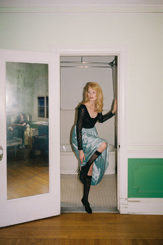 Woman in a black top and green skirt standing in a doorway with a room in the background.