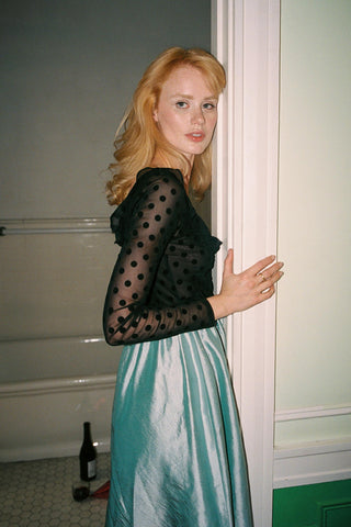Woman in a black polka dot top and green skirt standing in a room with a bottle on the floor.