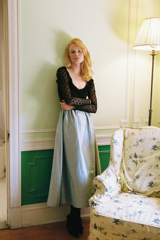Woman standing in a room with a floral-patterned chair and lamp.