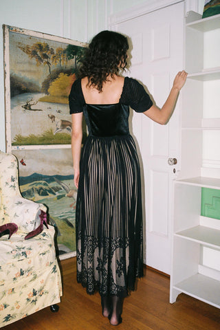 Woman in a black dress standing in a room with a painting and shelves.