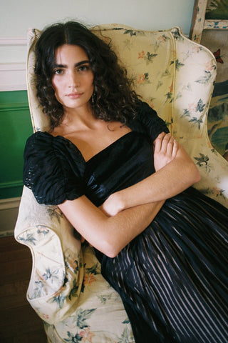 Woman in a black off-shoulder top and pleated skirt sitting on a floral-patterned chair.