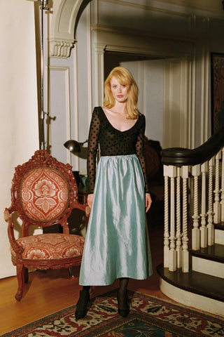 Woman in a black top and light green skirt standing in an elegant interior setting.