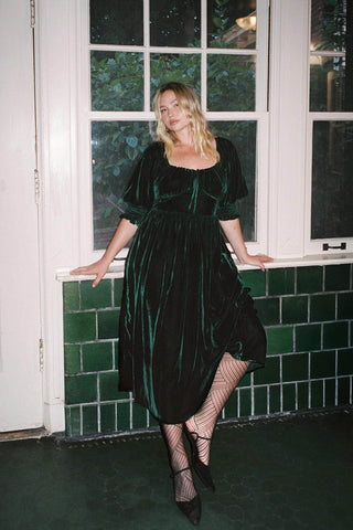 Woman in a green velvet dress standing in front of a window with green tiled walls.