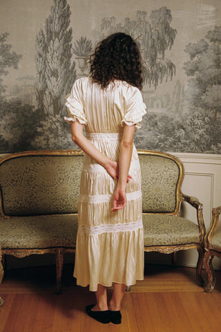 Woman in a vintage-style dress standing in a room with ornate wallpaper and furniture.