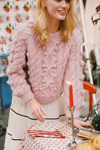 Woman wearing a rose colored cable knit sweater leaning over a table and candle