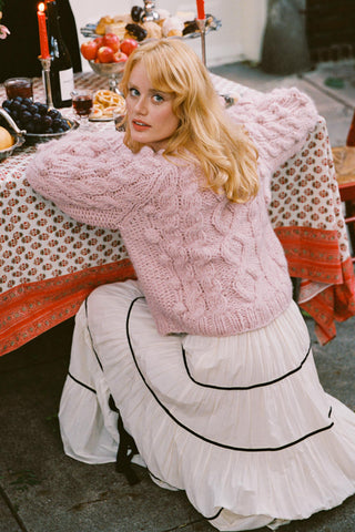 Woman wearing a rose colored cable knit sweater crouching in front of and leaning on a table