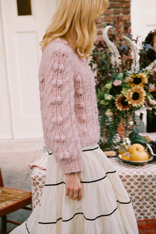 Side shot of a woman walking in a rose colored cable knit sweater and white skirt