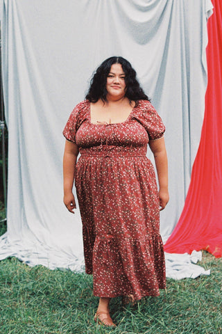 Woman wearing a red floral dress standing outdoors with a blue and red curtain in the background