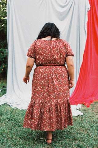 A back shot of a woman wearing a red floral dress standing outdoors with a blue and red curtain in the background
