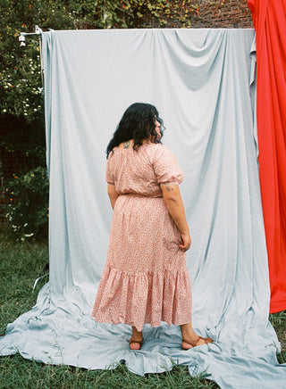 Back shot of a woman standing outdoors wearing a red floral dress with a blue curtain in the background