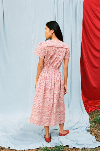 Woman in a red floral dress standing with her back to us against a light blue curtain with a red fabric partially visible.