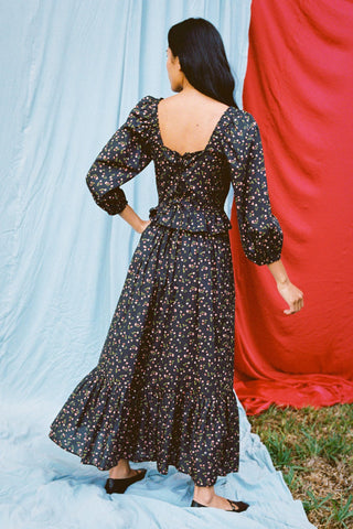 Back shot of a Woman wearing a black floral dress standing against a red and blue curtain.