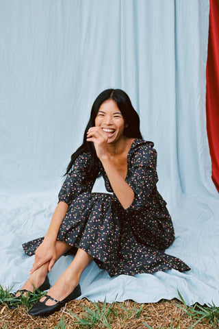 Woman in a black floral dress sitting on grass with a light blue curtain background