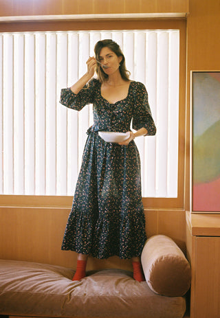 Woman in a floral dress standing on a couch eating in a room with a window and furniture.