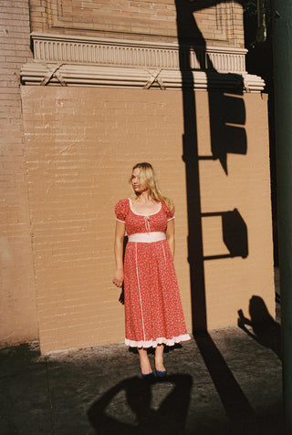 Woman wearing a red ground dress with white florals, a ribbon tie at chest, black flats leaning against a brick wall