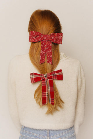 Person with red hair styled in two braids, each adorned with a different patterned bow, wearing a white sweater and blue jeans.