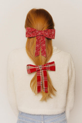 Person with red hair styled in two ponytails, each adorned with a red bow, against a plain background, one plaid, one red ribbon.