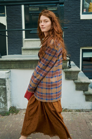 A woman with long red hair stands on a brick sidewalk in front of a dark brick building, wearing a brown and blue plaid blazer and a brown skirt.