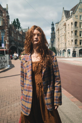 A woman with long, wavy red hair stands on a city street wearing a brown dress under a brown, blue, and beige plaid blazer.