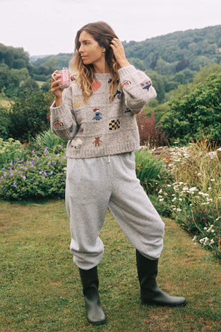 Woman in a garden wearing a patterned sweater and gray sweatpants, holding a pink mug.