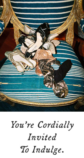 A pile of assorted women's shoes, including black heels, beige flats, and metallic silver pumps, resting on a blue and white striped upholstered armchair with gold trim. A white banner at the bottom contains the text 'You're Cordially Invited To Indulge.