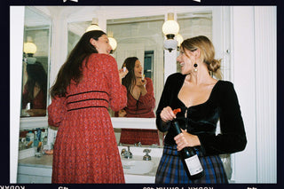 Two women standing in front of a bathroom mirror, one wearing a red patterned dress and the other in a black velvet top and plaid skirt while holding a bottle. The photo has a vintage, film-photo style with visible borders and lighting.