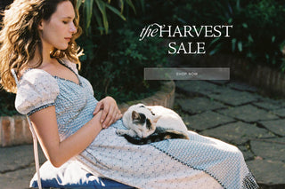 A woman in a light blue and white patterned dress sits outdoors with a cat resting on her lap, with the text 'the HARVEST SALE' and a 'SHOP NOW' button overlaid on the scene.