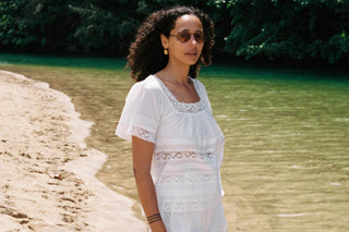 Woman in a white dress standing on a beach with water and greenery in the background
