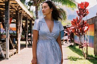 A woman wearing a light blue patterned short-sleeve wrap dress walks outdoors in a tropical setting with palm trees and colorful plants.