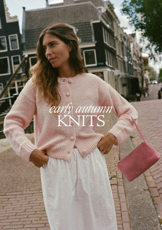 A woman standing on a cobblestone street in front of traditional Dutch houses, wearing a light pink knit cardigan with buttons and a white skirt, holding a pink handbag. The text 'early autumn KNITS' is overlaid across the center.