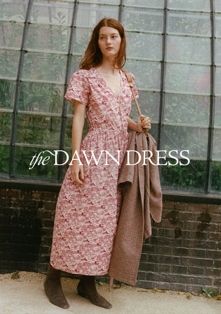 A woman stands outdoors in front of a greenhouse-style glass wall, wearing a pink floral midi dress with short sleeves and a V-neck. She holds a brown plaid coat and carries a shoulder bag.