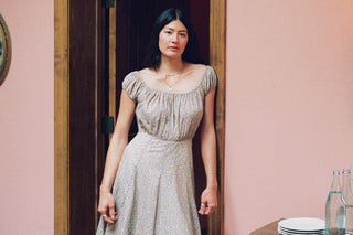 A woman stands in a doorway wearing a light beige textured dress with a square neckline and short ruffled sleeves, set against a soft pink wall.